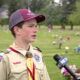 Boy scout in uniform being interviewed by a reporter in a grassy park filled with small flower decorations in the background.