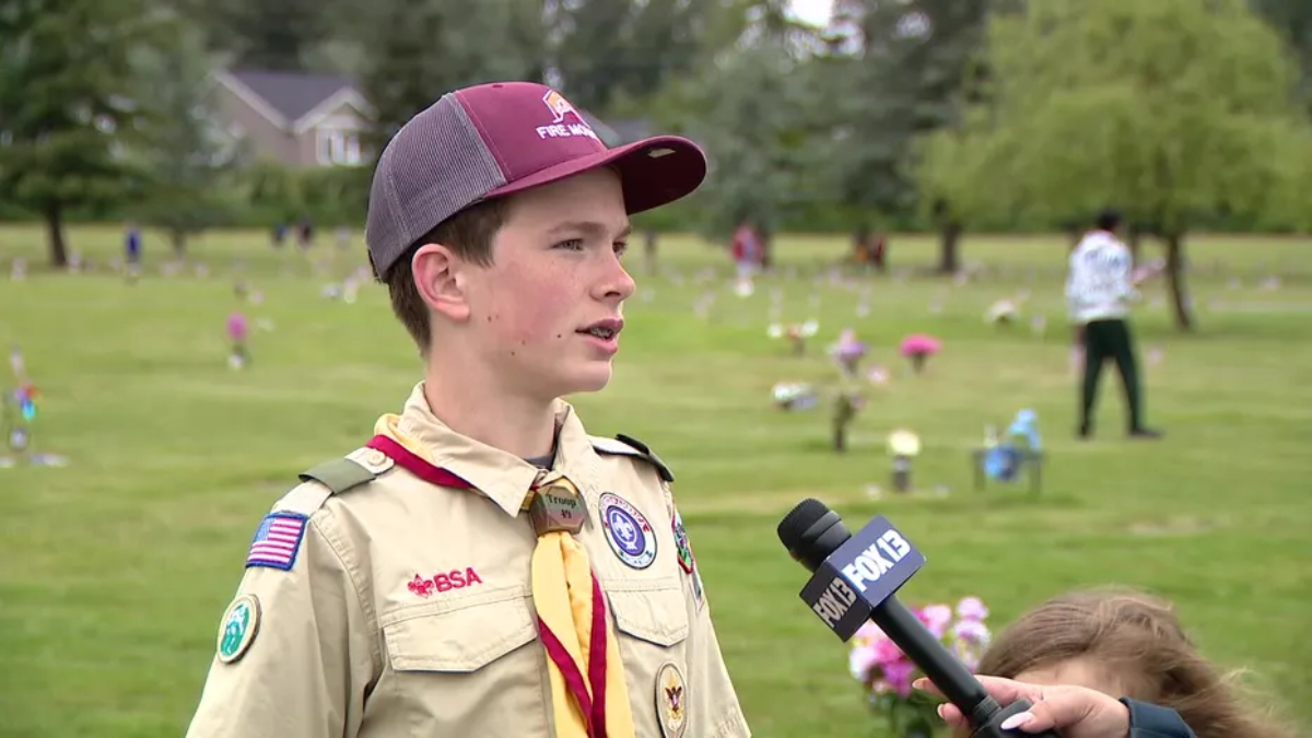 Boy scout in uniform being interviewed by a reporter in a grassy park filled with small flower decorations in the background.