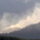 Large plume of wildfire smoke sweeping over a forested mountain slope in Oregon, with dense pine trees at the foreground and a bright, smoky sky above.