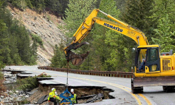Yellow Komatsu excavator hoists equipment to repair a collapsed mountain road with two workers in safety gear nearby.