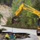 Yellow Komatsu excavator hoists equipment to repair a collapsed mountain road with two workers in safety gear nearby.