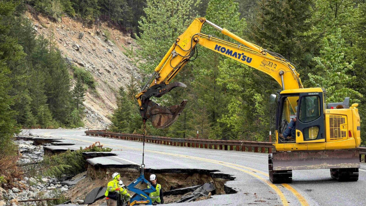Yellow Komatsu excavator hoists equipment to repair a collapsed mountain road with two workers in safety gear nearby.