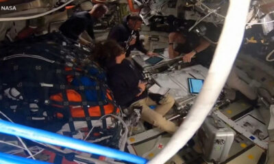 Astronauts seated and working around a central console in a cramped spacecraft cabin, with tablets and equipment visible and a NASA label in the corner.