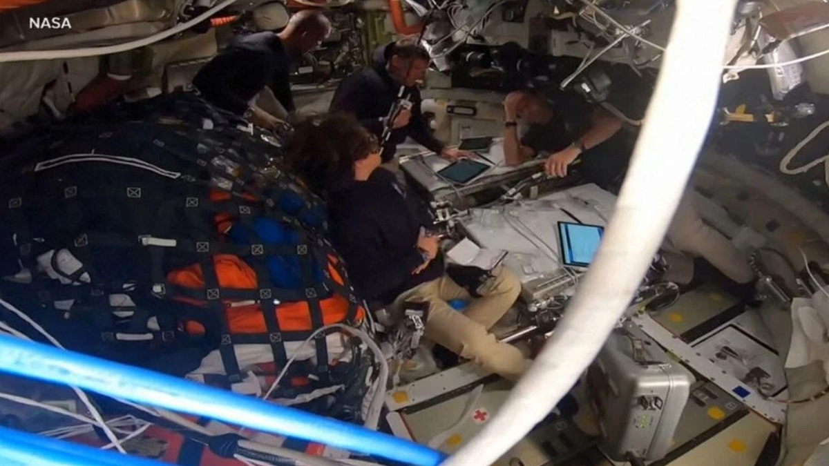 Astronauts seated and working around a central console in a cramped spacecraft cabin, with tablets and equipment visible and a NASA label in the corner.