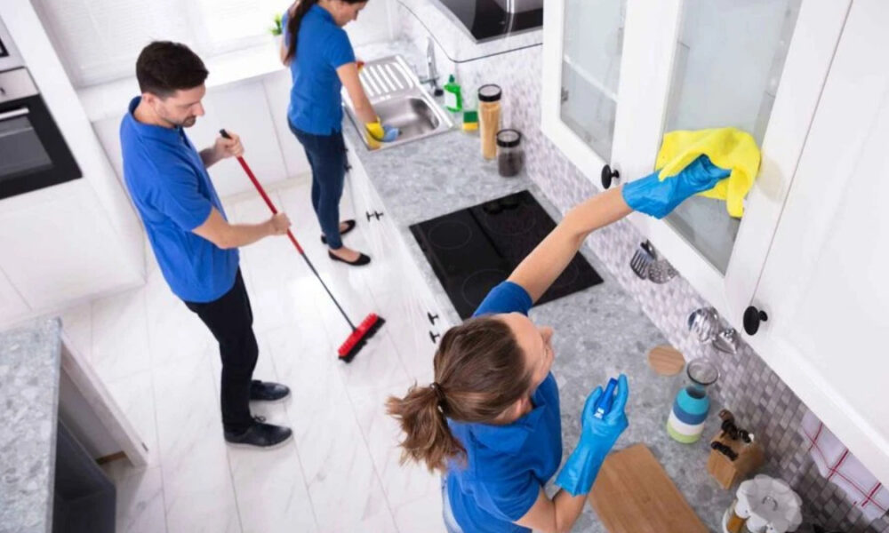 Three people in blue uniforms cleaning a bright white kitchen; one mops the floor while another wipes a window with a yellow cloth, and a third cleans the sink area.
