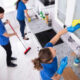 Three people in blue uniforms cleaning a bright white kitchen; one mops the floor while another wipes a window with a yellow cloth, and a third cleans the sink area.
