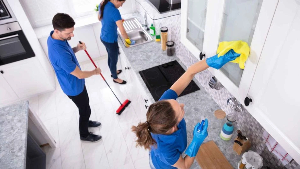 Three people in blue uniforms cleaning a bright white kitchen; one mops the floor while another wipes a window with a yellow cloth, and a third cleans the sink area.