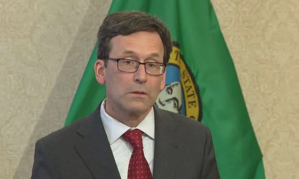 Man in a dark suit and red tie wearing glasses, standing in front of a green flag with a seal.