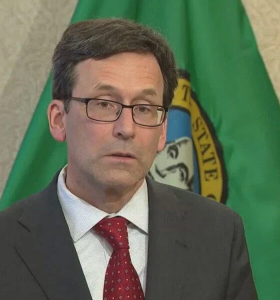 Man in a dark suit and red tie wearing glasses, standing in front of a green flag with a seal.