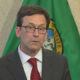 Man in a dark suit and red tie wearing glasses, standing in front of a green flag with a seal.