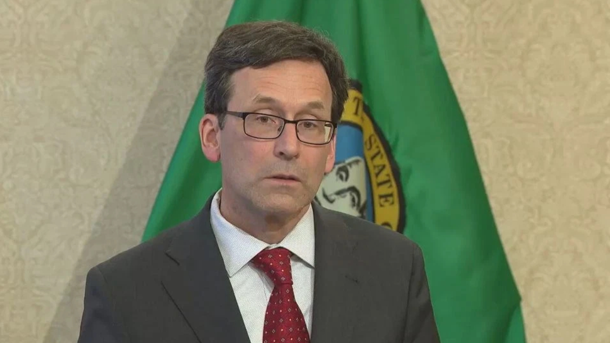Man in a dark suit and red tie wearing glasses, standing in front of a green flag with a seal.