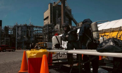 Hazmat-suited workers set up equipment at an industrial plant with orange traffic cones nearby.