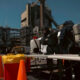 Hazmat-suited workers set up equipment at an industrial plant with orange traffic cones nearby.