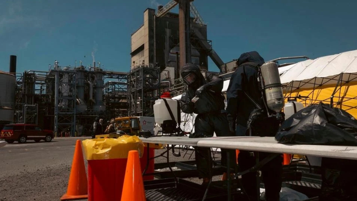 Hazmat-suited workers set up equipment at an industrial plant with orange traffic cones nearby.