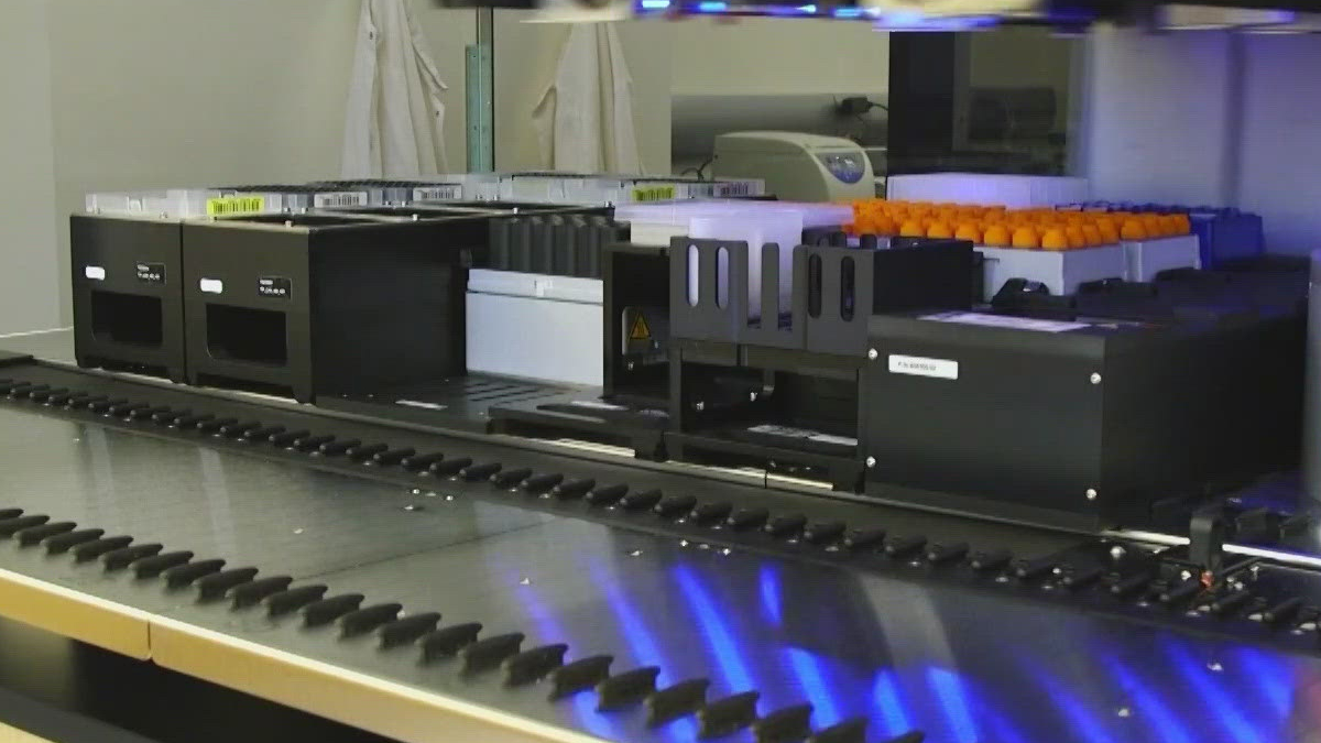 Laboratory analyzer with orange-capped tubes on a rack and a conveyor belt system under blue lights