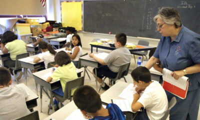 A teacher stands beside students at their desks, helping them with worksheets in a sunny classroom.