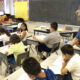 A teacher stands beside students at their desks, helping them with worksheets in a sunny classroom.