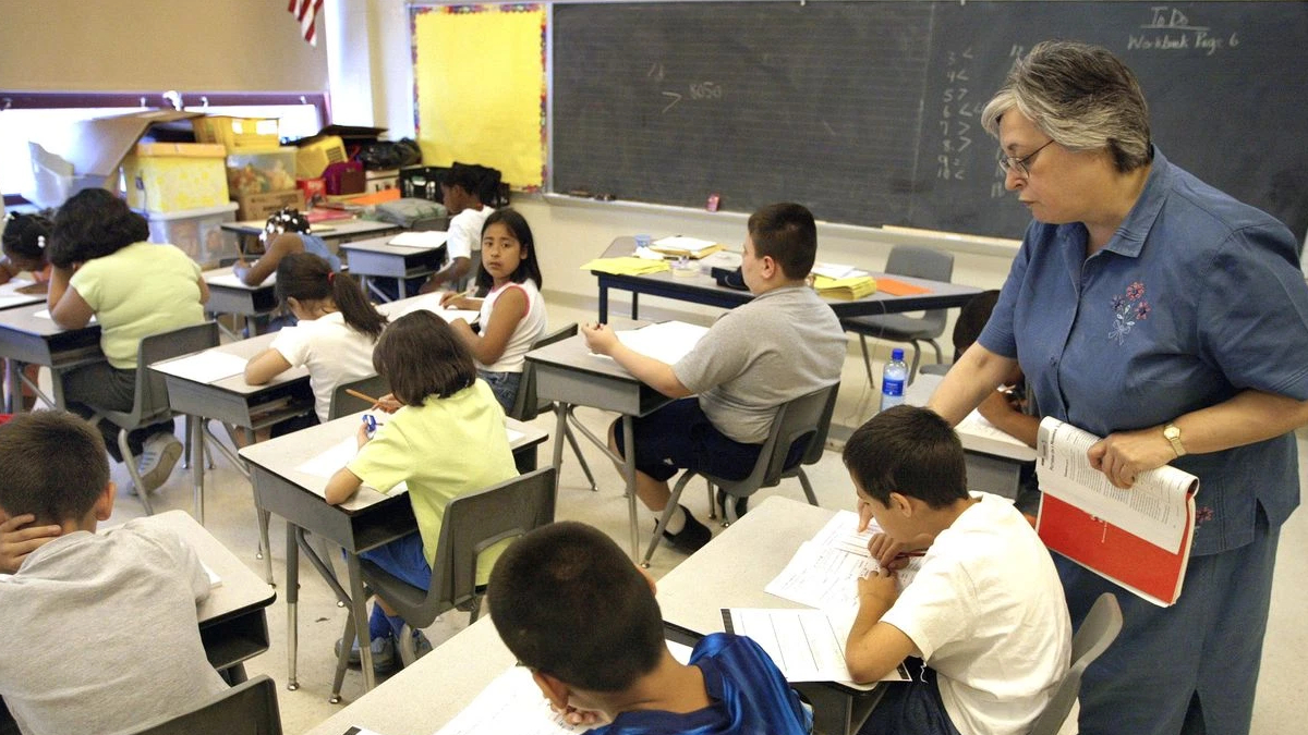 A teacher stands beside students at their desks, helping them with worksheets in a sunny classroom.