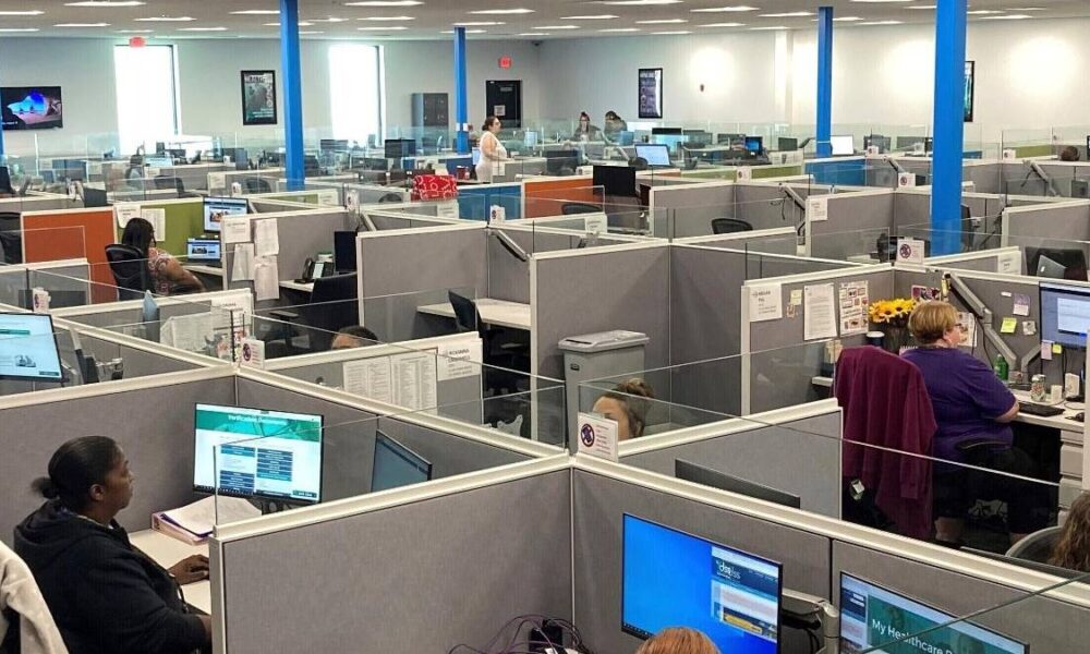 Open-plan call center with gray cubicles, blue support columns, and staff working at computer desks.