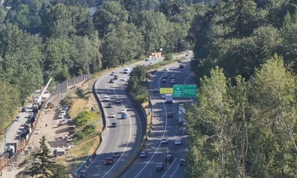 Aerial view of a multi-lane highway winding through a forest, with construction fencing and equipment on the left side far from the road edge.