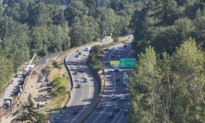Aerial view of a multi-lane highway winding through a forest, with construction fencing and equipment on the left side far from the road edge.