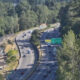 Aerial view of a multi-lane highway winding through a forest, with construction fencing and equipment on the left side far from the road edge.