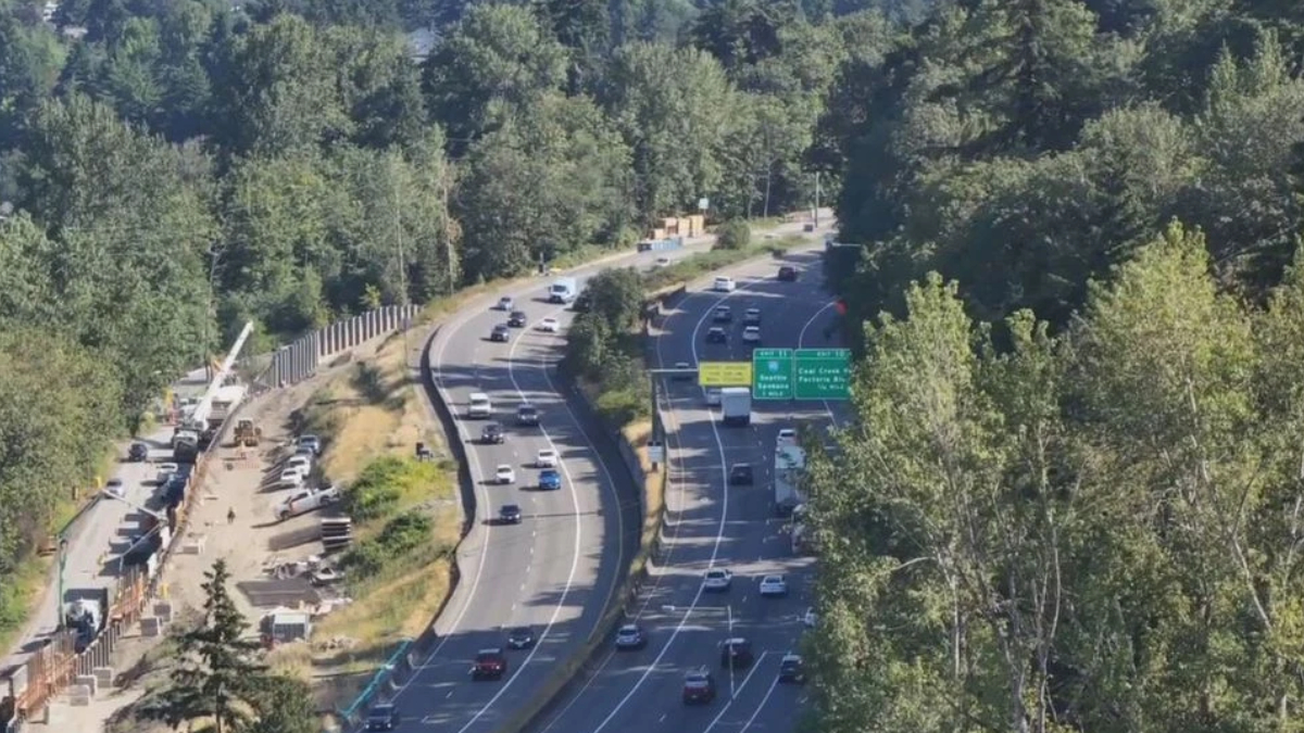 Aerial view of a multi-lane highway winding through a forest, with construction fencing and equipment on the left side far from the road edge.