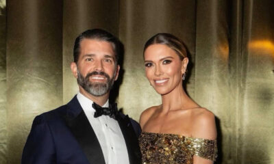 A well-dressed couple posing for a photo at a formal event; man in a navy tuxedo and bow tie, woman in a gold sequin off-shoulder gown, both smiling.