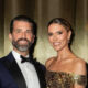 A well-dressed couple posing for a photo at a formal event; man in a navy tuxedo and bow tie, woman in a gold sequin off-shoulder gown, both smiling.