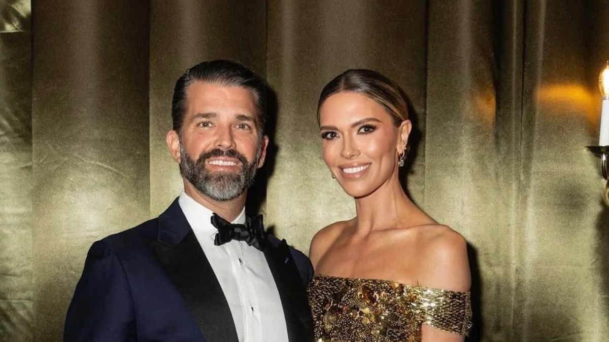 A well-dressed couple posing for a photo at a formal event; man in a navy tuxedo and bow tie, woman in a gold sequin off-shoulder gown, both smiling.