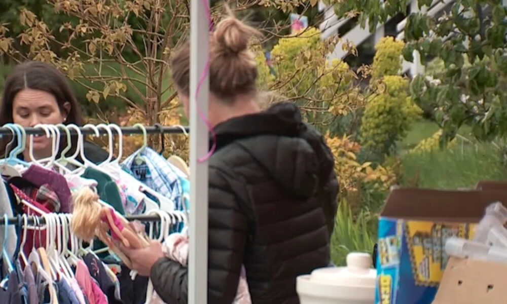 Woman in a black puffer jacket browses a rack of children's clothes at an outdoor sale, holding a doll. (informative)