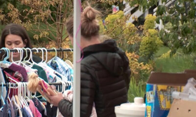 Woman in a black puffer jacket browses a rack of children's clothes at an outdoor sale, holding a doll. (informative)