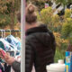 Woman in a black puffer jacket browses a rack of children's clothes at an outdoor sale, holding a doll. (informative)