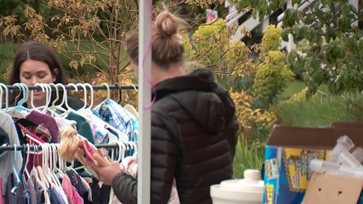 Woman in a black puffer jacket browses a rack of children's clothes at an outdoor sale, holding a doll. (informative)
