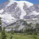 Snow-capped mountain rising above a green forest and meadow under a blue sky.