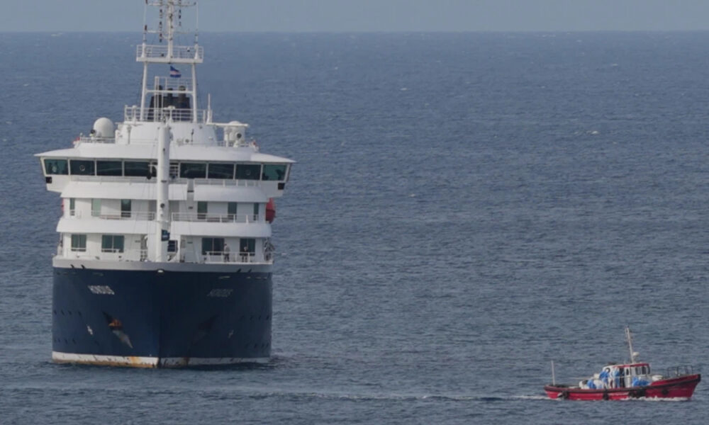 Front view of a large white and blue passenger ship on calm ocean, with a small red motorboat sailing nearby behind it.