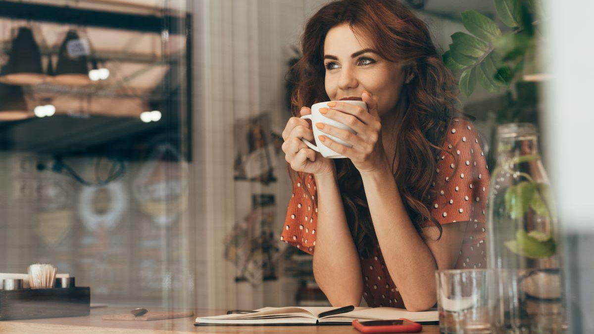Woman in a red polka-dot blouse sipping coffee at a cafe table, with an open notebook nearby.