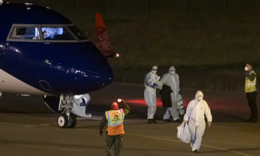 Night-time scene at an airport: a blue-and-white aircraft on the tarmac with crew in protective hazmat suits unloading luggage and assisting passengers.