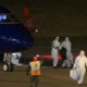 Night-time scene at an airport: a blue-and-white aircraft on the tarmac with crew in protective hazmat suits unloading luggage and assisting passengers.