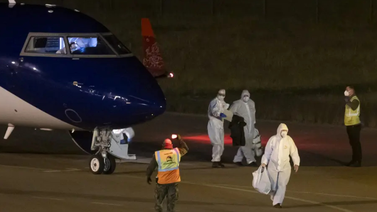 Night-time scene at an airport: a blue-and-white aircraft on the tarmac with crew in protective hazmat suits unloading luggage and assisting passengers.