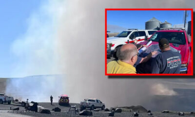 Wide view of a wildfire with thick smoke, emergency vehicles, and security cones on a barren ground. Inset shows firefighters reviewing a map near a red pickup truck, framed in red.