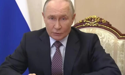 A man in a dark suit speaks at a desk in a formal setting, with a blue flag behind him and an ornate chair nearby.