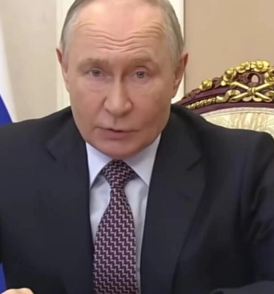 A man in a dark suit speaks at a desk in a formal setting, with a blue flag behind him and an ornate chair nearby.