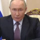 A man in a dark suit speaks at a desk in a formal setting, with a blue flag behind him and an ornate chair nearby.