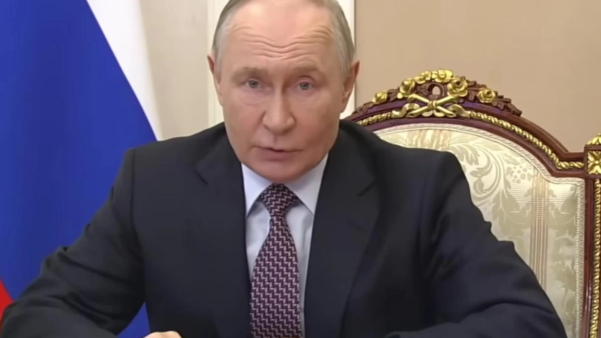A man in a dark suit speaks at a desk in a formal setting, with a blue flag behind him and an ornate chair nearby.