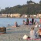 Crowded city beach along a calm bay with sunbathers, families, and swimmers; buildings and hills in the background.