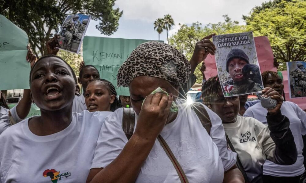 Crowd of protesters in white shirts, a woman in the foreground wipes away a tear while holding signs and photos demanding justice for David Kuloba.