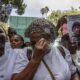 Crowd of protesters in white shirts, a woman in the foreground wipes away a tear while holding signs and photos demanding justice for David Kuloba.