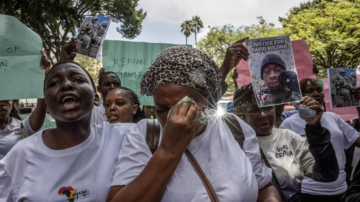 Crowd of protesters in white shirts, a woman in the foreground wipes away a tear while holding signs and photos demanding justice for David Kuloba.