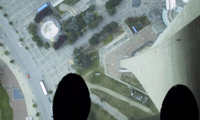 Aerial view from a high ledge showing a city square, roads, and a circular plaza, with dark shoes visible at the bottom edge as if standing on a tall structure.